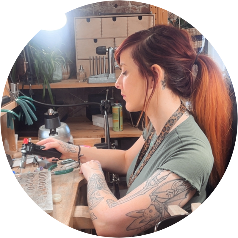 Image of Carley Pearson, a young woman with red hair tied back in a ponytail. She wears a green t-shirt and has tattoos on the arm closest to the camera. She is sat at a workbench, holding a a blowtorch over a number of metal rings laid on a ceramic plate.