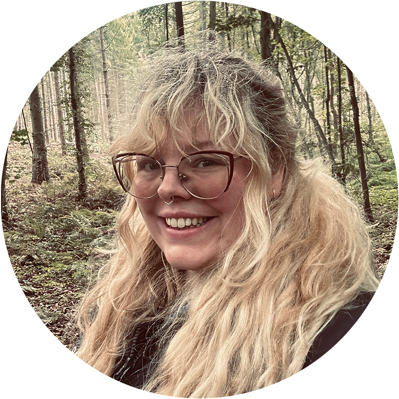 Image shows Jessica Leigh. A young woman with long blond hair. She is wearing rimmed glasses and a black jacket. She is standing with woodland in the background.